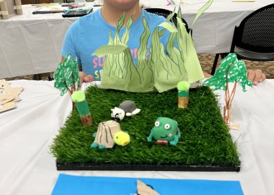 A girl seated at a table with green grass, featuring a clay frog, paper turtle, and clay snail nearby.