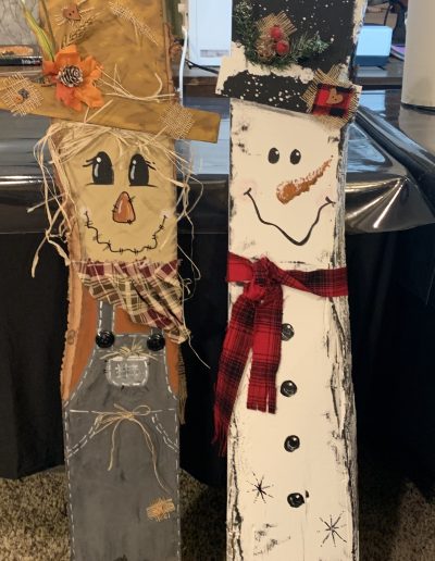 A scarecrow and snowman wearing hats and scarves, crafted from wood boards, stand together in a winter setting.