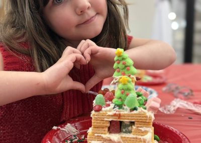 Child in red making a heart shape with hands next to a decorated gingerbread house on a red plate.