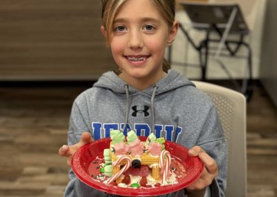 A person smiling and holding a red plate with a decorated gingerbread house.