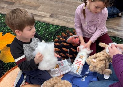 Two children crafting teddy bears with stuffing on a colorful rug.