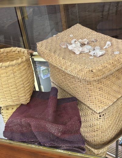 Woven baskets displayed in a glass case, with seashells arranged on the lid of a stacked basket and a burgundy textile underneath.
